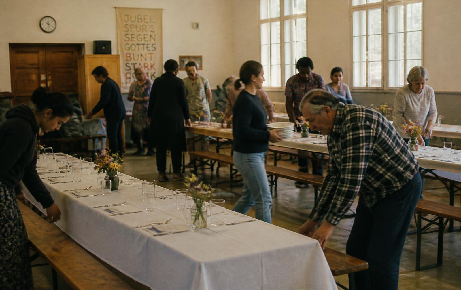 Neighbors of varied ages working together to set long trestle tables for a shared community meal in a plain hall